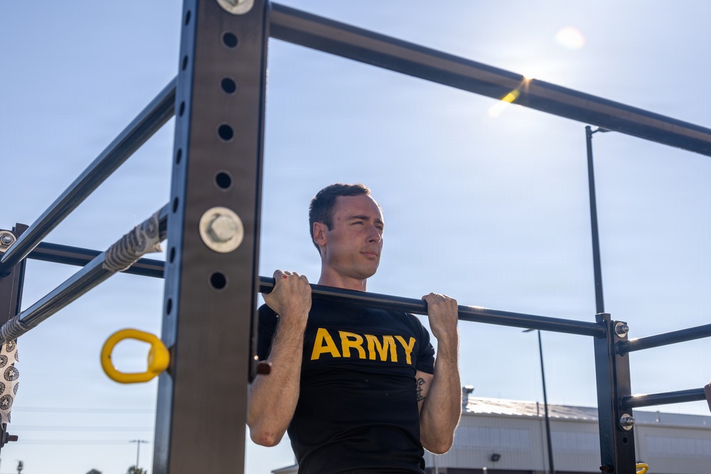 A young male soldier wearing a black U.S. Army PT (physical training) shirt is performing pull-ups on an outdoor military fitness station. The image is taken from a low angle looking upward, with bright sunlight visible in the background against a clear blue sky. The setting appears to be an outdoor exercise area, likely on a military installation.