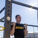 A young male soldier wearing a black U.S. Army PT (physical training) shirt is performing pull-ups on an outdoor military fitness station. The image is taken from a low angle looking upward, with bright sunlight visible in the background against a clear blue sky. The setting appears to be an outdoor exercise area, likely on a military installation.