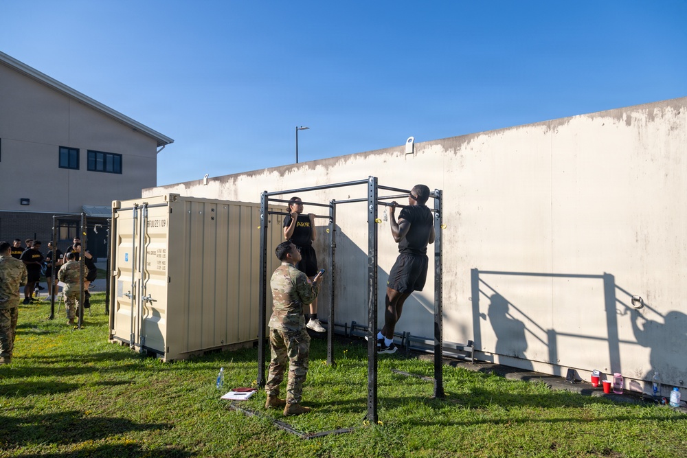 Military personnel are conducting a physical fitness test or training exercise outdoors on a grassy area adjacent to a large concrete wall. A soldier in camouflage uniform appears to be evaluating or counting repetitions as another soldier performs pull-ups on a metal pull-up bar station. Additional personnel in Army PT (physical training) uniforms are visible in the background engaged in similar activities.