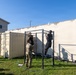 Military personnel are conducting a physical fitness test or training exercise outdoors on a grassy area adjacent to a large concrete wall. A soldier in camouflage uniform appears to be evaluating or counting repetitions as another soldier performs pull-ups on a metal pull-up bar station. Additional personnel in Army PT (physical training) uniforms are visible in the background engaged in similar activities.