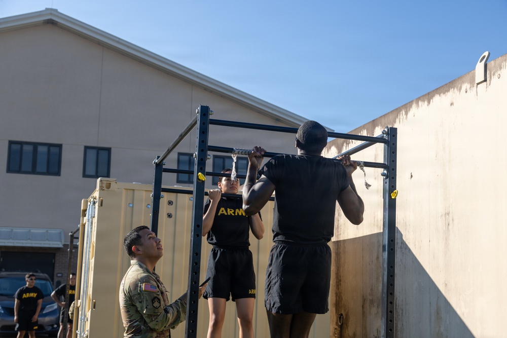Two U.S. Army soldiers in physical training (PT) uniforms are performing pull-ups on an outdoor pull-up bar rack. A third soldier in Army Combat Uniform (ACU) is observing and likely grading or supervising the exercise. The setting appears to be a military installation or training area.