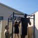Two U.S. Army soldiers in physical training (PT) uniforms are performing pull-ups on an outdoor pull-up bar rack. A third soldier in Army Combat Uniform (ACU) is observing and likely grading or supervising the exercise. The setting appears to be a military installation or training area.