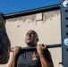 A bald man is performing pull-ups on an outdoor exercise bar, visibly exerting effort with a strained expression. He is wearing a dark gray t-shirt with a military-style unit patch/emblem on the chest. The photo is taken from a low angle against a blue sky and a concrete wall background.