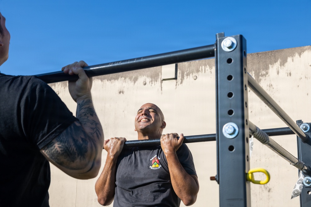 Two individuals are performing pull-ups on an outdoor metal pull-up bar rack against a concrete wall backdrop under a clear blue sky. The central figure, a bald man wearing a dark t-shirt with a visible emblem, is mid-exercise with his chin near the bar and an expression of exertion. A second person on the left, partially visible, has a tattooed arm and is also gripping the same bar.