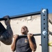 Two individuals are performing pull-ups on an outdoor metal pull-up bar rack against a concrete wall backdrop under a clear blue sky. The central figure, a bald man wearing a dark t-shirt with a visible emblem, is mid-exercise with his chin near the bar and an expression of exertion. A second person on the left, partially visible, has a tattooed arm and is also gripping the same bar.