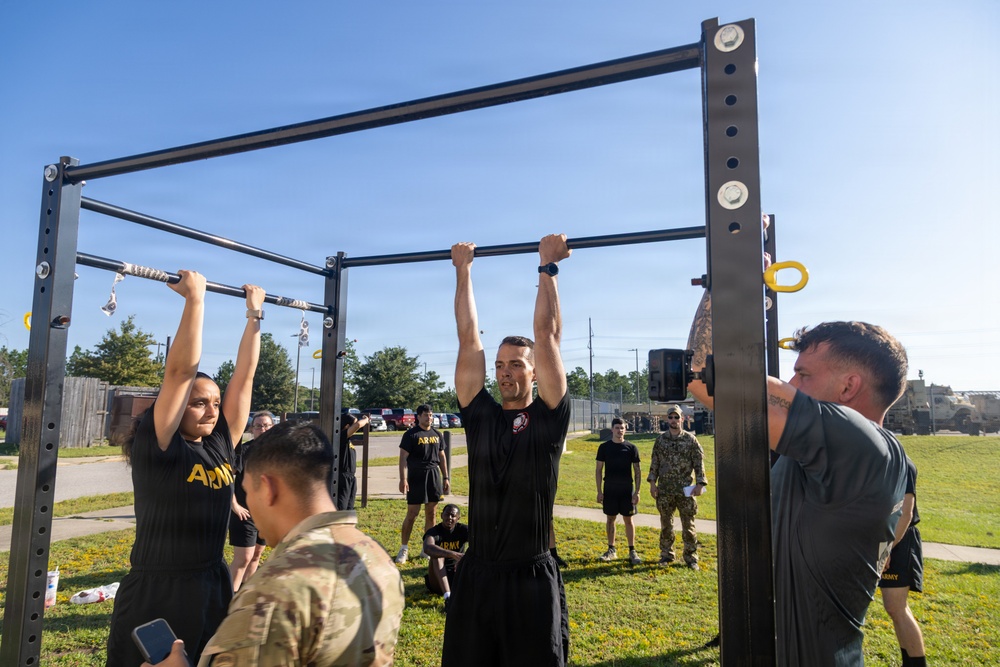 U.S. Army soldiers are performing pull-ups on an outdoor fitness rack during what appears to be a physical fitness test or training exercise. Two soldiers — one female and one male — are actively doing pull-ups while observers and evaluators watch nearby. The setting is an outdoor military installation on a clear, sunny day.