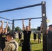 U.S. Army soldiers are performing pull-ups on an outdoor fitness rack during what appears to be a physical fitness test or training exercise. Two soldiers — one female and one male — are actively doing pull-ups while observers and evaluators watch nearby. The setting is an outdoor military installation on a clear, sunny day.