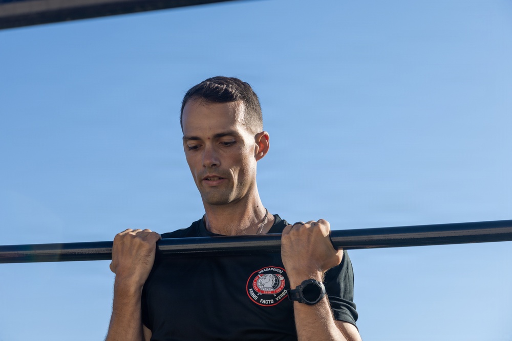 A lean, short-haired man is performing pull-ups on a horizontal bar outdoors against a clear blue sky. He is wearing a black t-shirt with a circular patch/emblem on the chest and a dark sports watch on his left wrist. His gaze is directed downward, suggesting focused physical exertion.