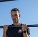 A lean, short-haired man is performing pull-ups on a horizontal bar outdoors against a clear blue sky. He is wearing a black t-shirt with a circular patch/emblem on the chest and a dark sports watch on his left wrist. His gaze is directed downward, suggesting focused physical exertion.
