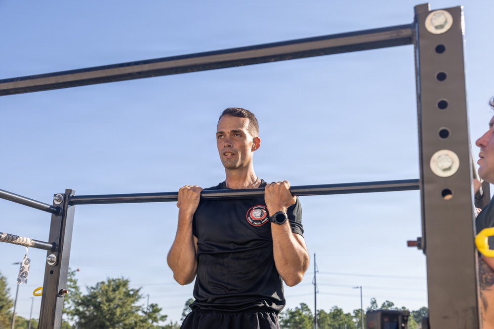 A young man in a black athletic t-shirt is performing pull-ups on an outdoor fitness bar structure. He appears to be at the top of the movement with his chin at bar level, wearing a watch on his left wrist. Another person is partially visible on the right side of the frame, suggesting this may be a supervised fitness test or training session.