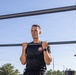 A young man in a black athletic t-shirt is performing pull-ups on an outdoor fitness bar structure. He appears to be at the top of the movement with his chin at bar level, wearing a watch on his left wrist. Another person is partially visible on the right side of the frame, suggesting this may be a supervised fitness test or training session.