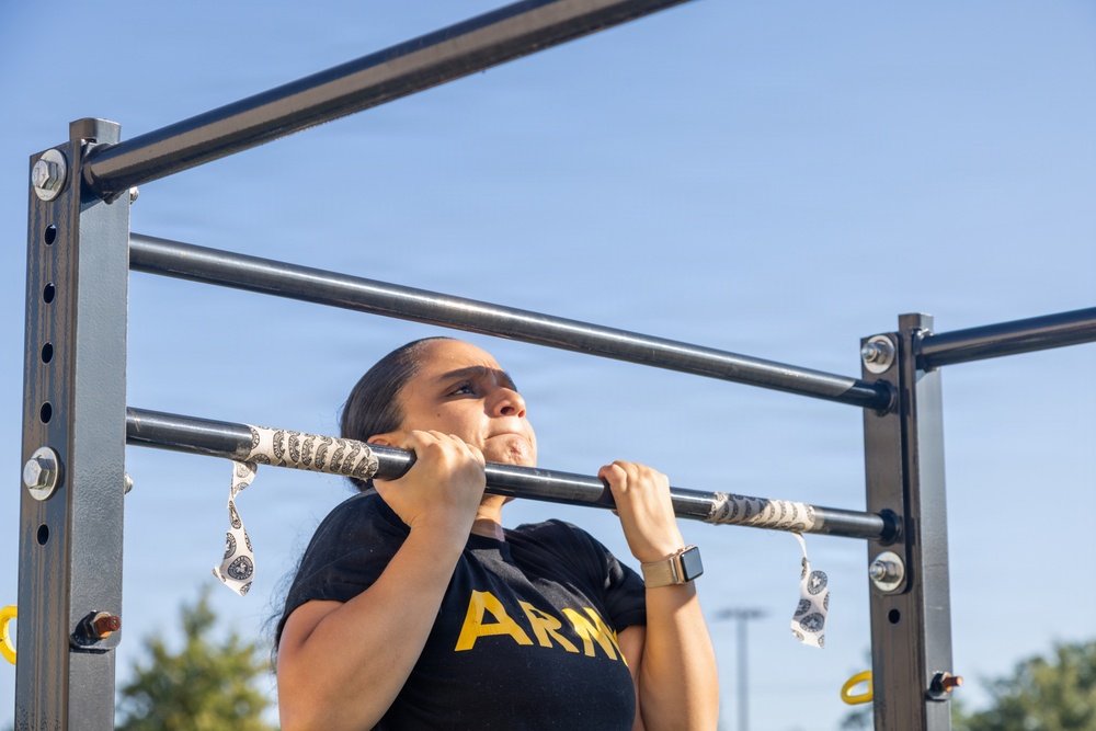 A woman wearing a black U.S. Army t-shirt is performing pull-ups on an outdoor fitness bar structure. She appears to be mid-exercise, with her chin at bar level, displaying visible physical exertion. The setting is an outdoor military or public fitness area under a clear blue sky.