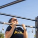A woman wearing a black U.S. Army t-shirt is performing pull-ups on an outdoor fitness bar structure. She appears to be mid-exercise, with her chin at bar level, displaying visible physical exertion. The setting is an outdoor military or public fitness area under a clear blue sky.