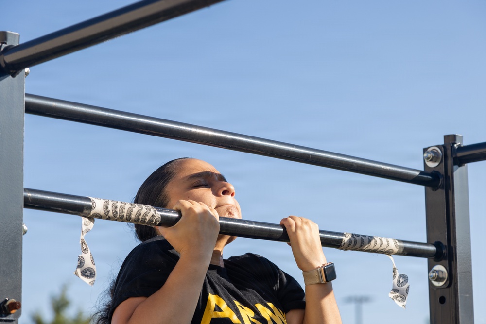 A woman is performing pull-ups on an outdoor exercise bar, with her chin raised above the bar indicating an active repetition. She is wearing a black t-shirt with 'ARMY' text visible, a smartwatch, and has her hair pulled back. The pull-up bar is wrapped with a paisley-patterned bandana for grip.
