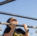 A woman is performing pull-ups on an outdoor exercise bar, with her chin raised above the bar indicating an active repetition. She is wearing a black t-shirt with 'ARMY' text visible, a smartwatch, and has her hair pulled back. The pull-up bar is wrapped with a paisley-patterned bandana for grip.
