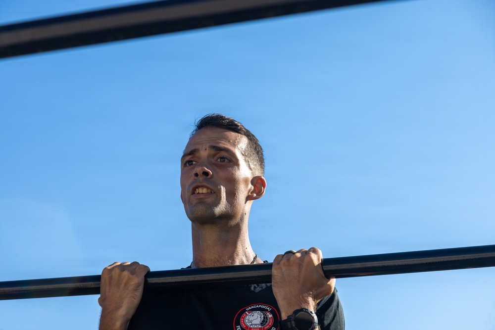 A man is performing pull-ups on an outdoor horizontal bar against a clear blue sky. He appears to be mid-exercise, with his chin near or above the bar, showing visible physical exertion. He is wearing a dark t-shirt with a circular patch/logo and a watch on his right wrist.