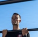 A man is performing pull-ups on an outdoor horizontal bar against a clear blue sky. He appears to be mid-exercise, with his chin near or above the bar, showing visible physical exertion. He is wearing a dark t-shirt with a circular patch/logo and a watch on his right wrist.