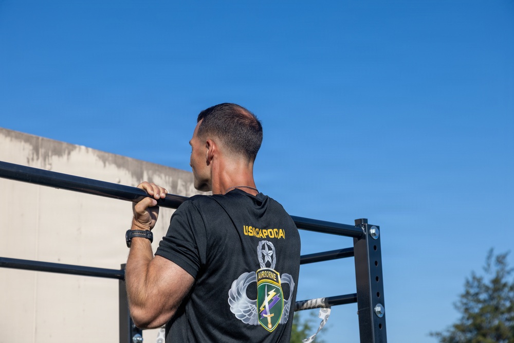 A muscular male soldier is performing pull-ups on an outdoor metal bar structure under a clear blue sky. He is wearing a black military t-shirt with 'USACAPOC(A)' and an Airborne insignia on the back. The setting appears to be a military fitness or training area.