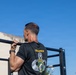 A muscular male soldier is performing pull-ups on an outdoor metal bar structure under a clear blue sky. He is wearing a black military t-shirt with 'USACAPOC(A)' and an Airborne insignia on the back. The setting appears to be a military fitness or training area.