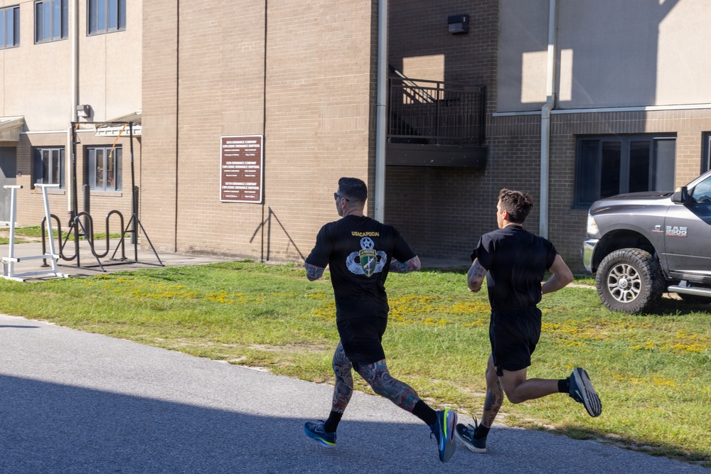 Two individuals wearing black athletic clothing are running along a paved path on what appears to be a military installation. The runner in the foreground is wearing a black t-shirt with an Airborne unit insignia and has heavily tattooed legs. A Ram 2500 pickup truck and military-style buildings with a unit sign board are visible in the background.