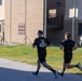 Two individuals wearing black athletic clothing are running along a paved path on what appears to be a military installation. The runner in the foreground is wearing a black t-shirt with an Airborne unit insignia and has heavily tattooed legs. A Ram 2500 pickup truck and military-style buildings with a unit sign board are visible in the background.