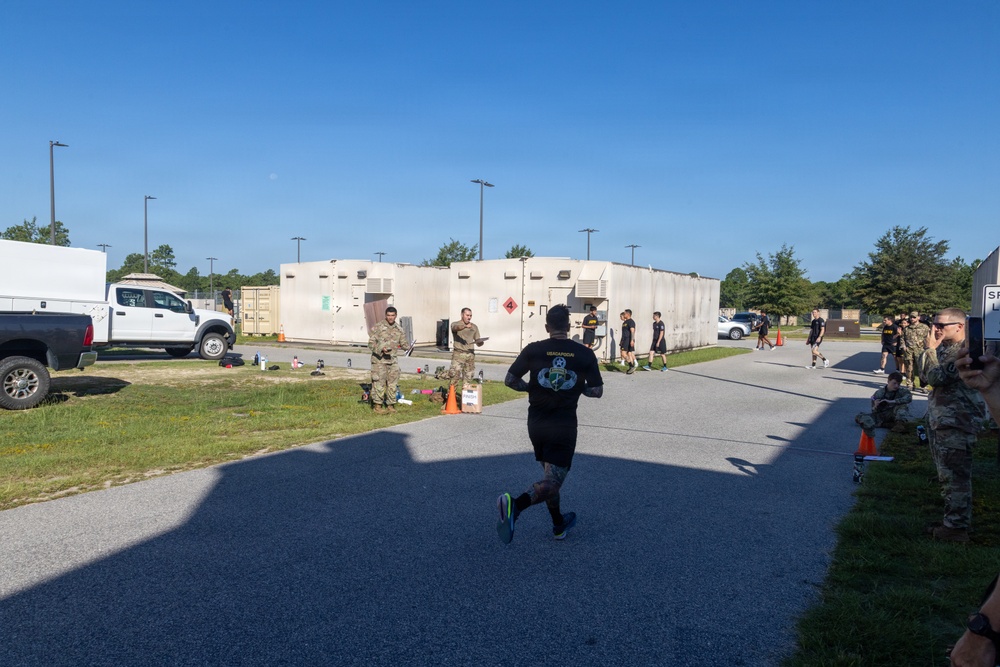 A military fitness event or physical training test is taking place on a paved area of what appears to be a military installation. A runner in black civilian athletic wear is approaching a finish or checkpoint area where uniformed military personnel are stationed. Observers in military uniforms line the right side of the course.