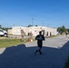 A military fitness event or physical training test is taking place on a paved area of what appears to be a military installation. A runner in black civilian athletic wear is approaching a finish or checkpoint area where uniformed military personnel are stationed. Observers in military uniforms line the right side of the course.