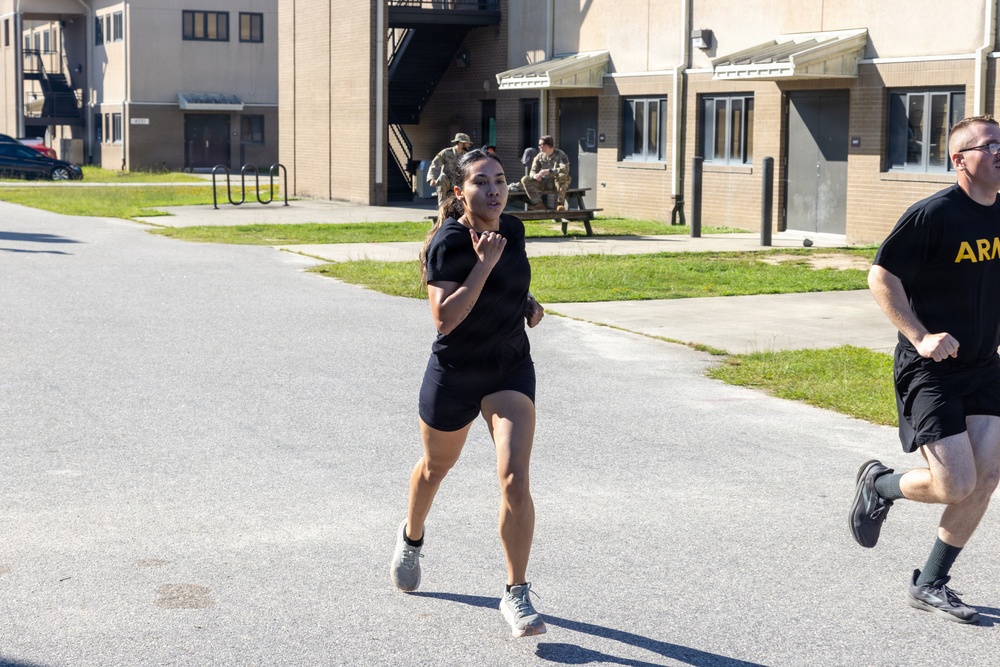 Two individuals in black athletic wear are running on a paved road at what appears to be a U.S. Army military installation. The person in the foreground is a woman running with focused expression, while a man wearing a shirt with 'ARMY' text is visible on the right edge. In the background, uniformed soldiers are seated at a picnic table near multi-story brick barracks buildings.