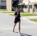 Two individuals in black athletic wear are running on a paved road at what appears to be a U.S. Army military installation. The person in the foreground is a woman running with focused expression, while a man wearing a shirt with 'ARMY' text is visible on the right edge. In the background, uniformed soldiers are seated at a picnic table near multi-story brick barracks buildings.