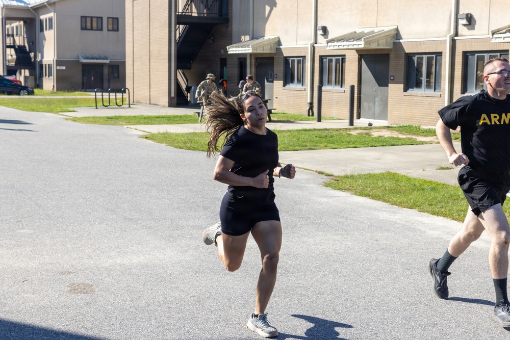 Two individuals in U.S. Army physical training (PT) uniforms are running on a paved road on what appears to be a military installation. A woman in a black t-shirt and shorts leads the run, with a man wearing an 'ARMY' branded black PT shirt visible behind her. Additional personnel in military uniforms can be seen in the background near brick barracks buildings.