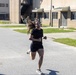 Two individuals in U.S. Army physical training (PT) uniforms are running on a paved road on what appears to be a military installation. A woman in a black t-shirt and shorts leads the run, with a man wearing an 'ARMY' branded black PT shirt visible behind her. Additional personnel in military uniforms can be seen in the background near brick barracks buildings.