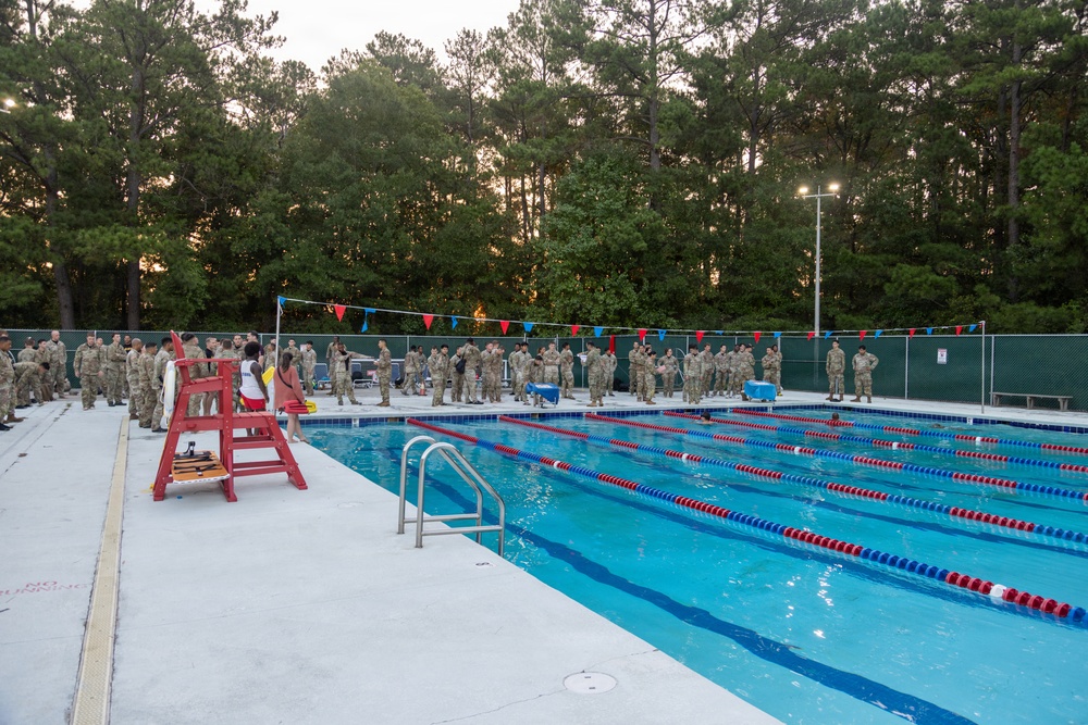 A large group of military personnel in camouflage uniforms are assembled around an outdoor swimming pool, likely at a military installation. The scene appears to be taking place at dusk or dawn, with a lifeguard chair visible on the pool deck and lane markers in the water. The gathering suggests a formal military swim qualification or water survival training event.