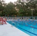 A large group of military personnel in camouflage uniforms are assembled around an outdoor swimming pool, likely at a military installation. The scene appears to be taking place at dusk or dawn, with a lifeguard chair visible on the pool deck and lane markers in the water. The gathering suggests a formal military swim qualification or water survival training event.