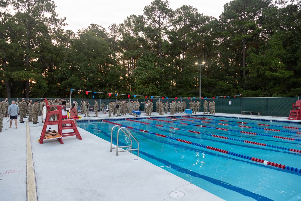 A large group of military personnel in camouflage uniforms are assembled on the far side of an outdoor competitive swimming pool, likely for a briefing or water training exercise. The pool features multiple lap lanes marked with red and blue lane dividers, and a lifeguard chair is visible in the foreground. The scene appears to be taking place at dusk or early morning at what appears to be a military installation.