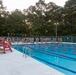 A large group of military personnel in camouflage uniforms are assembled on the far side of an outdoor competitive swimming pool, likely for a briefing or water training exercise. The pool features multiple lap lanes marked with red and blue lane dividers, and a lifeguard chair is visible in the foreground. The scene appears to be taking place at dusk or early morning at what appears to be a military installation.