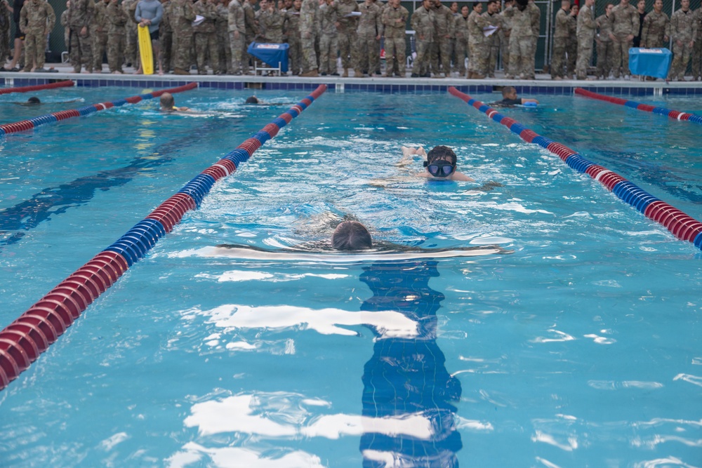Military personnel are conducting a swimming exercise or assessment in an indoor pool with red and blue lane dividers. A large group of soldiers in camouflage uniforms observes from the poolside. At least two swimmers are visible in the lanes, with one wearing a diving mask.