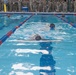 Military personnel are conducting a swimming exercise or assessment in an indoor pool with red and blue lane dividers. A large group of soldiers in camouflage uniforms observes from the poolside. At least two swimmers are visible in the lanes, with one wearing a diving mask.