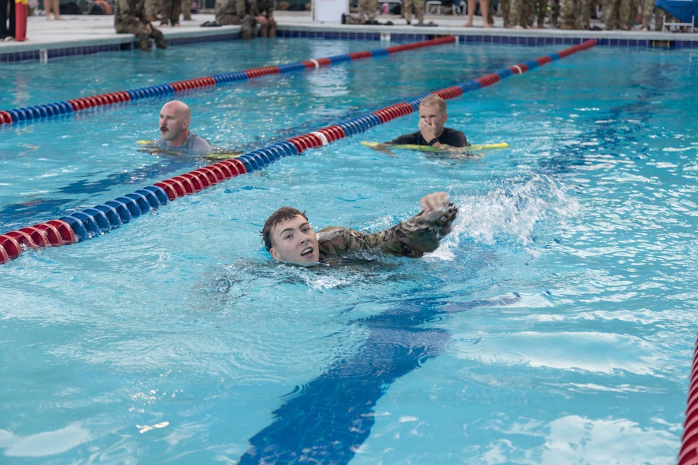Military personnel are conducting a swimming exercise in an indoor competition pool, wearing camouflage uniforms while swimming. A young soldier in the foreground is performing a freestyle stroke, while two observers or instructors in the background monitor from the water using kickboards. A crowd of uniformed personnel watches from the pool deck in the background.