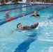 Military personnel are conducting a swimming exercise in an indoor competition pool, wearing camouflage uniforms while swimming. A young soldier in the foreground is performing a freestyle stroke, while two observers or instructors in the background monitor from the water using kickboards. A crowd of uniformed personnel watches from the pool deck in the background.