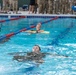 Military personnel are conducting a combat water survival training exercise in an indoor swimming pool. Soldiers in full camouflage uniforms are swimming laps while a safety observer monitors from the water. A large group of uniformed soldiers observes from the pool deck in the background.