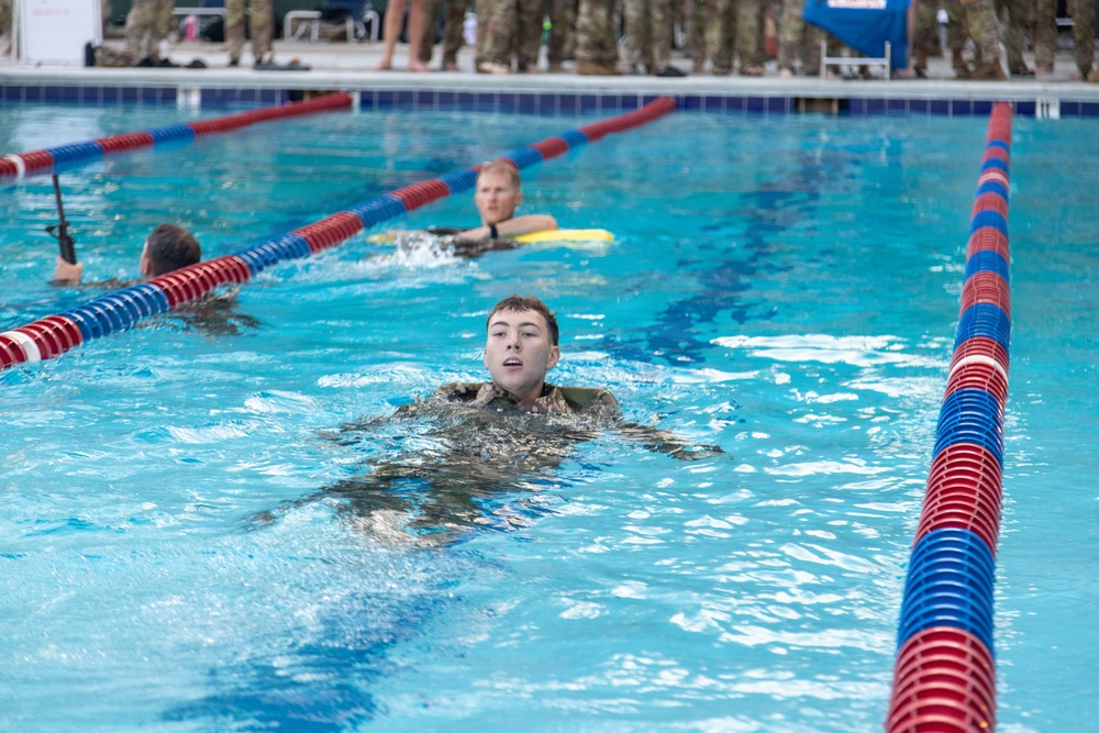 Military personnel are swimming in full camouflage uniforms in a competition or training pool with red and blue lane dividers. At least three swimmers are visible in separate lanes, with one swimmer prominently in the foreground. A large group of uniformed observers watches from the pool deck in the background.