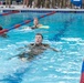 Military personnel are swimming in full camouflage uniforms in a competition or training pool with red and blue lane dividers. At least three swimmers are visible in separate lanes, with one swimmer prominently in the foreground. A large group of uniformed observers watches from the pool deck in the background.