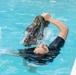 A person is floating on their back in a clear blue swimming pool, holding what appears to be wet camouflage clothing or fabric raised above the water. The individual is wearing a black outfit and appears to be in motion, with water splashing around them. The pool edge is visible on the right side of the frame.