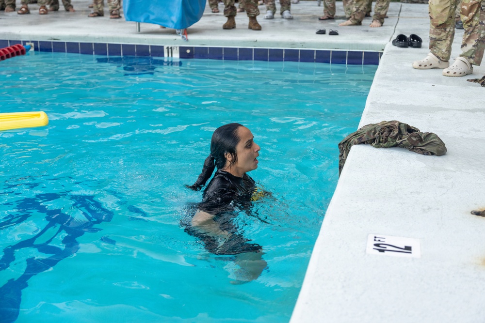 A person in wet black clothing is in a swimming pool, appearing to be participating in a military water survival or swim qualification training exercise. Multiple uniformed military personnel are visible standing around the pool deck observing. A wet camouflage uniform is draped over the pool edge, suggesting the swimmer recently removed it.