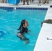 A person in wet black clothing is in a swimming pool, appearing to be participating in a military water survival or swim qualification training exercise. Multiple uniformed military personnel are visible standing around the pool deck observing. A wet camouflage uniform is draped over the pool edge, suggesting the swimmer recently removed it.