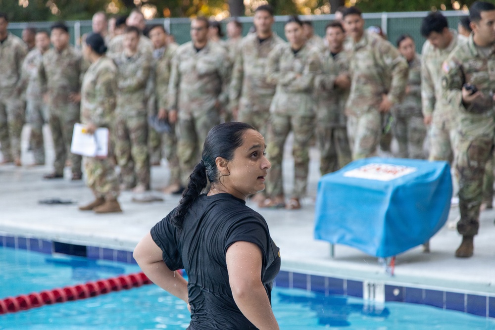 A woman in wet black clothing stands at the edge of a swimming pool, appearing to have just completed a swim. Behind her, a large group of military personnel in camouflage uniforms (OCP/MultiCam pattern) observe from poolside. The setting appears to be a military swim qualification or water survival training event.