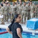 A woman in wet black clothing stands at the edge of a swimming pool, appearing to have just completed a swim. Behind her, a large group of military personnel in camouflage uniforms (OCP/MultiCam pattern) observe from poolside. The setting appears to be a military swim qualification or water survival training event.