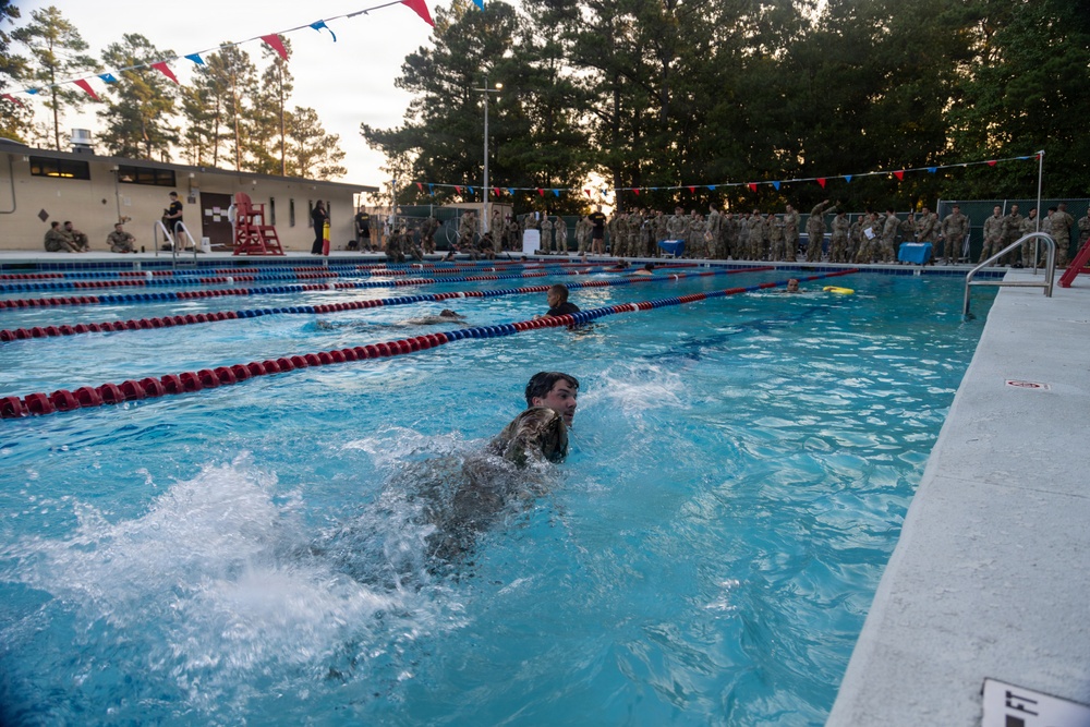 Military personnel in combat uniforms are conducting a swimming assessment or competition in an outdoor pool facility. A large group of uniformed soldiers lines the pool deck observing the swimmers, while several individuals actively swim laps. The setting appears to be a military installation pool during early morning or evening hours based on the lighting conditions.