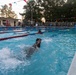 Military personnel in combat uniforms are conducting a swimming assessment or competition in an outdoor pool facility. A large group of uniformed soldiers lines the pool deck observing the swimmers, while several individuals actively swim laps. The setting appears to be a military installation pool during early morning or evening hours based on the lighting conditions.