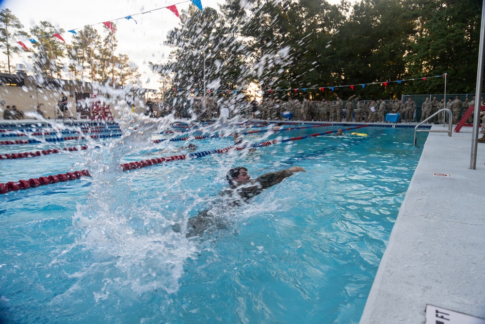 A swimmer in what appears to be military uniform or clothing is actively swimming in an outdoor competition pool, generating a large water splash in the foreground. A crowd of individuals dressed in military camouflage uniforms lines the poolside in the background, suggesting this is a military athletic event or competition. The scene is decorated with red, white, and blue pennant flags, indicating a formal or celebratory occasion.